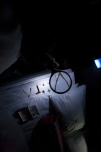 MATTHEW SWARTS A DIMLY LIT, CONTEMPORARY SCENE SHOWS A WHITE BED WITH OBJECTS INCLUDING A BOOK, PLIERS, SMALL FIGURINES, AND A MAGNIFYING GLASS HIGHLIGHTING A WISHBONE—EVOKING THE MOOD OF AN INTRIGUING VISUAL OR VIDEO INSTALLATION. A PILLOW AND STRIPED BAG ARE ALSO VISIBLE.