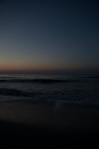 MATTHEW SWARTS A DIMLY LIT BEACH AT DUSK WITH GENTLE OCEAN WAVES, A DARK SHORELINE, AND A FAINT ORANGE AND BLUE GRADIENT IN THE SKY AS THE SUN SETS.