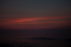 MATTHEW SWARTS A DARK, TRANQUIL OCEAN IS BARELY VISIBLE UNDER A DUSKY SKY, ILLUMINATED BY SOFT RED AND PINK HUES FROM THE SETTING OR RISING SUN. THE SCENE IS PEACEFUL WITH GENTLE WAVES AND A SUBTLE GRADIENT OF COLORS.