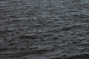 MATTHEW SWARTS DARK, RIPPLING WATER WITH SMALL WAVES COVERING THE SURFACE, CREATING A TEXTURED PATTERN. THE SCENE SUGGESTS A BODY OF WATER UNDER OVERCAST OR LOW LIGHT CONDITIONS. NO LAND, SKY, OR OTHER OBJECTS ARE VISIBLE.
