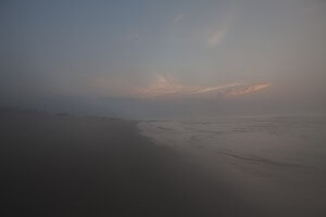 MATTHEW SWARTS A MISTY BEACH AT DUSK WITH GENTLE WAVES, DARK SAND, AND SOFT PINK CLOUDS ILLUMINATED BY THE SETTING SUN IN A HAZY SKY.