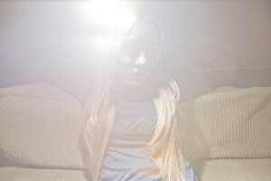 MATTHEW SWARTS A PERSON WITH LONG BLONDE HAIR SITS ON A BEIGE COUCH, PARTIALLY OBSCURED BY A BRIGHT LIGHT FLARE. THE HAZY, ETHEREAL EFFECT EVOKES CONTEMPORARY ART PHOTOGRAPHY OR AN ABSTRACT VIDEO STILL.