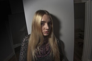 MATTHEW SWARTS A YOUNG WOMAN WITH LONG BLONDE HAIR, WEARING A DARK LACE TOP WITH A RUFFLED COLLAR, STANDS IN A DIMLY LIT ROOM. HER SERIOUS EXPRESSION AND THE VISUAL CONTRAST CREATE A STRIKING, CONTEMPORARY ATMOSPHERE.