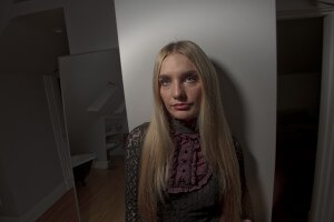 MATTHEW SWARTS A VISUAL, CONTEMPORARY PORTRAIT: A WOMAN WITH LONG BLONDE HAIR IN A DARK LACE DRESS WITH A HIGH-NECK, RUFFLED PURPLE COLLAR STANDS INDOORS BY A MIRROR, EVOKING THE DISTINCT STYLE OF MATTHEW SWARTS.