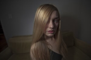 MATTHEW SWARTS A WOMAN WITH LONG, STRAIGHT BLONDE HAIR SITS ON A BEIGE SOFA IN A DIMLY LIT ROOM, LOOKING SLIGHTLY TO THE SIDE WITH A NEUTRAL EXPRESSION. THE SUBDUED LIGHTING AND BACKGROUND CREATE A CONTEMPORARY VISUAL ATMOSPHERE.