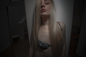 MATTHEW SWARTS A PERSON WITH LONG BLONDE HAIR STANDS AGAINST A WALL, WEARING A BLACK AND BLUE LACE BRA. THEIR FACE IS PARTIALLY COVERED BY HAIR, AND THE DRAMATIC LIGHTING CREATES A SHADOWY EFFECT, GIVING THE SCENE A STRIKING CONTEMPORARY VISUAL ART VIBE.