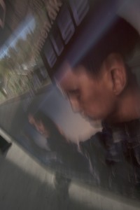 MATTHEW SWARTS A BLURRY PHOTO SHOWS A REFLECTION OF A PERSON’S FACE AND UPPER BODY IN A GLASS SURFACE, WITH PARTIALLY VISIBLE WHITE TEXT AND OUTDOOR SCENERY FAINTLY SEEN IN THE BACKGROUND—EVOKING THE SUBTLE CREATIVITY OF NORDIC ART FROM DENMARK OR NORWAY.