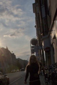 MATTHEW SWARTS A STREET SCENE AT SUNSET IN OSLO SHOWS A SILHOUETTED PERSON WALKING ON THE SIDEWALK, WITH PARKED BICYCLES, STREET SIGNS, AND BUILDINGS ON THE RIGHT. CARS DRIVE ALONG THE ROAD AS ARTFUL GOLDEN LIGHT CASTS LONG SHADOWS ACROSS THE CITYSCAPE.