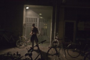 MATTHEW SWARTS A PERSON STANDS IN FRONT OF A LIT DOORWAY AT NIGHT IN DENMARK, SURROUNDED BY SEVERAL BICYCLES. THE BLURRY, DIM IMAGE FEATURES OUT-OF-FOCUS SHADOWS AND FAINT LIGHT HIGHLIGHTING THE FIGURE AND BIKES.