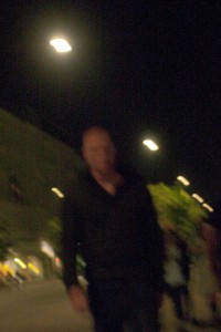 MATTHEW SWARTS A BLURRY PHOTO OF A PERSON STANDING OUTSIDE AT NIGHT UNDER STREETLIGHTS IN OSLO, WITH TREES AND BUILDINGS VISIBLE IN THE BACKGROUND. THE IMAGE IS OUT OF FOCUS AND DIMLY LIT.