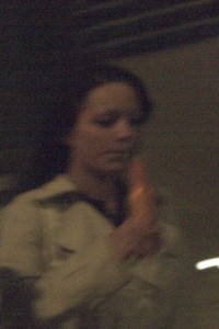 MATTHEW SWARTS A BLURRY, LOW-LIGHT IMAGE OF A PERSON IN A LIGHT-COLORED OUTFIT HOLDING AN ORANGE OBJECT, POSSIBLY A CARROT, EVOKES THE ENIGMATIC ART OF MATTHEW SWARTS, WITH INDISTINCT BACKGROUND DETAILS REMINISCENT OF AN OSLO EVENING.