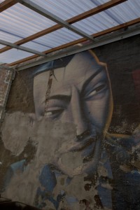 MATTHEW SWARTS A LARGE, WEATHERED MURAL OF A MAN'S FACE WITH AN INTENSE EXPRESSION IS PAINTED ON A BRICK WALL UNDER A TRANSLUCENT ROOF IN OSLO. PORTIONS OF THE MURAL ARE FADED AND PEELING, REVEALING THE ROUGH SURFACE BENEATH.