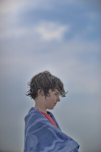 MATTHEW SWARTS A CHILD WITH WET, CURLY HAIR WRAPPED IN A BLUE TOWEL OVER A RED SHIRT, SEEN IN PROFILE AGAINST A SOFT BLUE CLOUDY SKY.