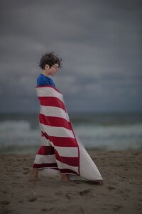 MATTHEW SWARTS A YOUNG BOY WRAPPED IN A RED AND WHITE STRIPED TOWEL WALKS ON A SANDY BEACH WITH A STORMY SKY AND OCEAN IN THE BACKGROUND.