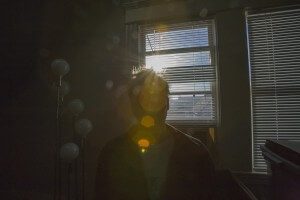 MATTHEW SWARTS A PERSON STANDS IN A DIMLY LIT ROOM WITH SUNLIGHT STREAMING THROUGH A WINDOW BEHIND THEM, CREATING LENS FLARES AND CASTING THEIR FIGURE IN SHADOW. VERTICAL BLINDS AND A MULTI-GLOBE FLOOR LAMP EVOKE THE MOODY STYLE OF MATTHEW SWARTS’ ART.