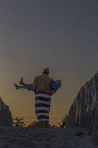 MATTHEW SWARTS A MAN IN AN ORANGE SHIRT AND STRIPED TOWEL CARRIES A LAUGHING CHILD ON A BEACH AT SUNSET.