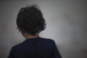 MATTHEW SWARTS A BLURRY, OUT-OF-FOCUS IMAGE SHOWING THE BACK OF A PERSONS HEAD WITH DARK, WAVY HAIR, WEARING A NAVY BLUE TOP, AGAINST A PLAIN GREY BACKGROUND.