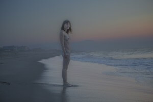 MATTHEW SWARTS A PERSON STANDS NUDE ON A BEACH AT DUSK, LOOKING TOWARD THE CAMERA. GENTLE WAVES WASH THE SHORE, AND THE SKY IS COLORED WITH SOFT HUES OF BLUE, PINK, AND PURPLE AS THE SUN SETS. HOUSES ARE VISIBLE IN THE DISTANT BACKGROUND.