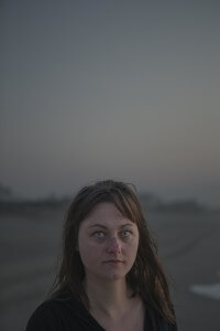 MATTHEW SWARTS CLOSE-UP PORTRAIT OF A WOMAN WITH BRIGHT BLUE EYES LOOKING AT THE CAMERA. THE BACKGROUND IS A BLURRY, HAZY LANDSCAPE.