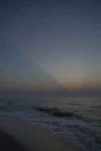 MATTHEW SWARTS A CALM OCEAN SHORELINE AT DUSK WITH SMALL WAVES GENTLY LAPPING THE SAND. THE SKY IS DARKENING, SHOWING SOFT GRADIENTS OF BLUE AND ORANGE, WITH A SUBTLE RAY OF LIGHT EXTENDING FROM THE HORIZON.