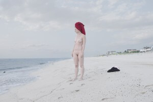 MATTHEW SWARTS A NUDE PERSON WITH A RED TOWEL WRAPPED AROUND THEIR HEAD STANDS ON A SANDY BEACH, GAZING TOWARD THE OCEAN. FOOTPRINTS TRAIL BEHIND, AND CLOTHES ARE LYING ON THE SAND NEARBY. THE SKY IS MOSTLY CLOUDY.