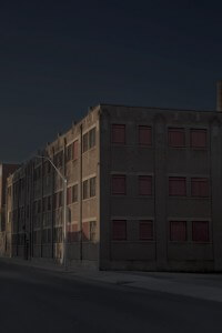 MATTHEW SWARTS AN IMPOSING, MULTI-STORY BRICK BUILDING WITH RED-COVERED WINDOWS STANDS AGAINST A DARK SKY. A STREET LAMP CASTS A LONG SHADOW ACROSS THE STREET IN FRONT OF IT.