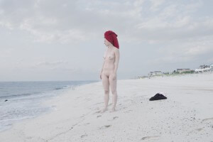 MATTHEW SWARTS A NUDE PALE-SKINNED WOMAN WITH A RED TOWEL WRAPPED AROUND HER HAIR STANDS ON A SANDY BEACH, GAZING TOWARD THE OCEAN. CLOTHES LIE ON THE SAND NEARBY. CLOUDY SKY AND DISTANT SHORELINE BUILDINGS VISIBLE.