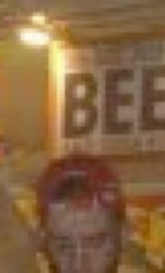 MATTHEW SWARTS A BLURRY IMAGE SHOWING THE TOP OF A PERSONS HEAD IN THE FOREGROUND AND A LARGE SIGN IN THE BACKGROUND WITH THE WORD BEER PARTIALLY VISIBLE. THE LIGHTING IS WARM AND THE SCENE APPEARS TO BE INDOORS.