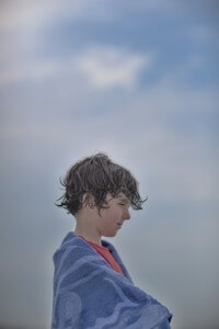MATTHEW SWARTS A CHILD WRAPPED IN A BLUE TOWEL LOOKS TO THE SIDE, WITH WET HAIR AND A RED SHIRT VISIBLE.