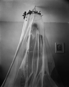 MATTHEW SWARTS A PERSON STANDS BEHIND A SHEER, DRAPED CURTAIN HANGING FROM A FLORAL HOOP ON THE CEILING IN A SOFTLY LIT ROOM. THEIR FORM IS BLURRED BY THE FABRIC, EVOKING CONTEMPORARY PORTRAITURE AS VISUAL ART. A FRAMED PICTURE HANGS ON THE WALL BEHIND THEM.