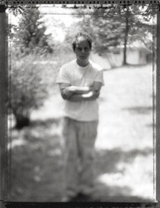 MATTHEW SWARTS BLACK AND WHITE PHOTO BY MATTHEW SWARTS OF A PERSON STANDING OUTDOORS WITH ARMS CROSSED, WEARING A WHITE SHIRT AND PANTS. THE BLURRY VISUAL FEATURES TREES AND A BUILDING IN THE BACKGROUND, ADDING AN ABSTRACT, DREAMLIKE QUALITY.