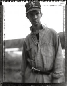 MATTHEW SWARTS BLACK AND WHITE PHOTOGRAPHY CAPTURES A MAN OUTDOORS IN A “HABITAT FOR HUMANITY” HAT, STRIPED SHIRT, AND BELT. AGAINST A BLURRY, NATURAL BACKGROUND, HE LOOKS DIRECTLY AT THE CAMERA WITH A NEUTRAL EXPRESSION, EMBODYING CONTEMPORARY PORTRAIT STYLE.