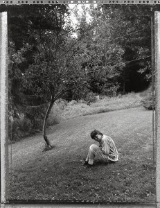 MATTHEW SWARTS A PERSON SITS ALONE ON A GRASSY SLOPE NEAR A SMALL TREE, THEIR KNEES DRAWN UP AND HEAD RESTING ON THEIR HANDS. THE BLACK AND WHITE IMAGE CAPTURES A QUIET, NATURAL LANDSCAPE IN A CONTEMPORARY DOCUMENTARY STYLE REMINISCENT OF MATTHEW SWARTS.