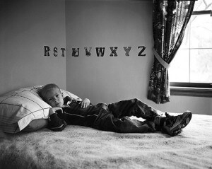 MATTHEW SWARTS A YOUNG BOY WITH CANCER LIES ON A BED, PROPPED UP ON A PILLOW, LOOKING THOUGHTFUL. BEHIND HIM, THE ALPHABET IS DISPLAYED ON THE WALL. SUNLIGHT STREAMS THROUGH A WINDOW WITH PATTERNED CURTAINS TO THE RIGHT.