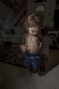 MATTHEW SWARTS A YOUNG, SHIRTLESS BOY WITH CURLY HAIR STANDS INDOORS ON A PATTERNED RUG, LOOKING DOWN. ANOTHER SMALL CHILD IS PARTLY VISIBLE IN THE BACKGROUND, BOTH INSIDE A COZY, DIMLY LIT LIVING ROOM—A CONTEMPORARY SCENE REMINISCENT OF MATTHEW SWARTS.
