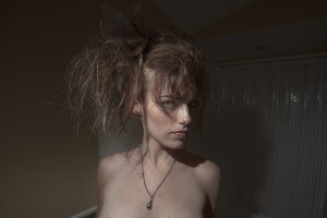 MATTHEW SWARTS A WOMAN WITH DRAMATIC, TEASED HAIR AND STRIKING BLUE EYES STANDS IN A DIMLY LIT ROOM, WEARING A NECKLACE. THE VISUAL ART SCENE FEATURES A WHITE SHOWER CURTAIN AND BEIGE WALLS IN THE BACKGROUND.