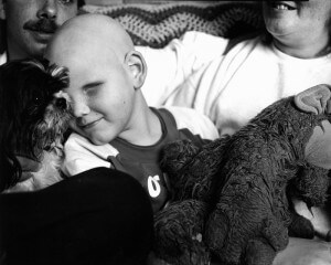 MATTHEW SWARTS A SMILING BALD CHILD, PART OF A CONTEMPORARY PORTRAITURE SERIES ON CHILDREN WITH CANCER, CUDDLES A FLUFFY DOG WHILE HOLDING A LARGE STUFFED ANIMAL. TWO ADULTS SIT NEARBY, THEIR WARMTH ADDING TO THIS TOUCHING PHOTOGRAPHY SCENE.
