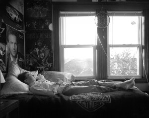 MATTHEW SWARTS A BOY LIES ON A BED COVERED WITH A HARLEY-DAVIDSON BLANKET IN A ROOM DECORATED WITH POSTERS OF CARS, A MUSICIAN, AND GUNS N ROSES. SUNLIGHT STREAMS THROUGH TWO WINDOWS ABOVE THE BED, ILLUMINATING THIS MOMENT OF PHOTOGRAPHY. A DREAMCATCHER HANGS BETWEEN THEM.