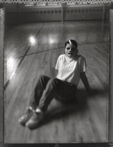 MATTHEW SWARTS A PERSON WEARING A MASK SITS ON THE FLOOR OF AN EMPTY GYMNASIUM WITH WOODEN FLOORING, LEGS EXTENDED AND ARMS BEHIND FOR SUPPORT—A CONTEMPORARY BLACK AND WHITE IMAGE BY MATTHEW SWARTS, SLIGHTLY OUT OF FOCUS.