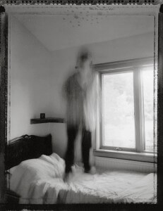 MATTHEW SWARTS A BLURRED, GHOSTLIKE FIGURE STANDS OR JUMPS ON A BED IN A SOFTLY LIT BEDROOM WITH A LARGE WINDOW AND MINIMAL DECOR, CREATING A SENSE OF MOTION IN THIS BLACK-AND-WHITE VISUAL REMINISCENT OF DOCUMENTARY PHOTOGRAPHY.