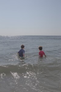 MATTHEW SWARTS TWO YOUNG BOYS STAND IN THE OCEAN WAVES, FACING AWAY FROM THE CAMERA. ONE WEARS A BLUE RASH GUARD, THE OTHER RED.