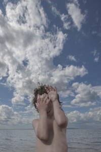 MATTHEW SWARTS A YOUNG PERSON COVERS THEIR FACE WITH THEIR HANDS AGAINST A BACKDROP OF A BLUE SKY FILLED WITH WHITE CLOUDS AND A CALM BODY OF WATER.
