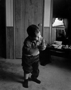 MATTHEW SWARTS A YOUNG CHILD STANDS INDOORS ON CARPET, HOLDING A TOY AND STUDYING IT CLOSELY. THIS INTIMATE SCENE, REMINISCENT OF CONTEMPORARY PORTRAITURE, FEATURES WOOD-PANELED WALLS, A CLOSED DOOR, AND TOYS AND DISHES SET OUT ON A TABLE NEARBY.