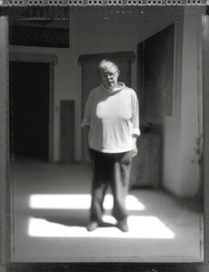 MATTHEW SWARTS A PERSON STANDS IN THE CENTER OF A SOFTLY LIT ROOM, FRAMED BY PATCHES OF LIGHT ON THE FLOOR. IN THIS MATTHEW SWARTS PHOTOGRAPH, THE BLACK AND WHITE IMAGE IS SLIGHTLY OUT OF FOCUS, CREATING AN ETHEREAL, DREAMLIKE EFFECT.