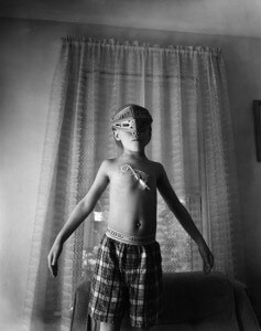 MATTHEW SWARTS BLACK AND WHITE PHOTO OF A SHIRTLESS BOY IN PLAID SHORTS WEARING A DECORATIVE MASK, ARMS OUTSTRETCHED, STANDING BEFORE SHEER CURTAINS IN A DIMLY LIT ROOM.