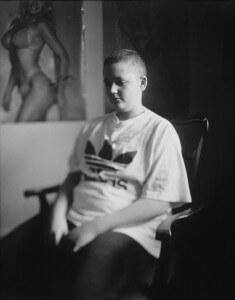 MATTHEW SWARTS A YOUNG PERSON WITH SHORT HAIR SITS ON A CHAIR IN A WHITE ADIDAS T-SHIRT. THIS BLACK AND WHITE PHOTOGRAPHY PIECE, SOFTLY FOCUSED, FEATURES A BLURRED POSTER OF A WOMAN IN A BIKINI ON THE WALL BEHIND—AN EXAMPLE OF CONTEMPORARY PORTRAITURE.