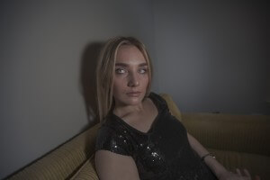 MATTHEW SWARTS A WOMAN WITH LONG BLONDE HAIR AND LIGHT EYES, WEARING A BLACK SEQUIN TOP, SITS ON A YELLOW COUCH AGAINST A GRAY WALL—CAPTURED IN CONTEMPORARY STYLE BY MATTHEW SWARTS—LOOKING SLIGHTLY PAST THE CAMERA WITH A SERIOUS EXPRESSION.