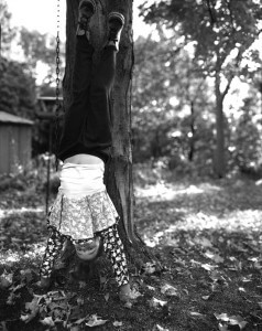 MATTHEW SWARTS A CHILD HANGS UPSIDE DOWN BY THEIR KNEES FROM A TREE BRANCH IN A SUNLIT YARD—A PLAYFUL MOMENT REMINISCENT OF MATTHEW SWARTS' PHOTOGRAPHY. SMILING, THE CHILD’S HANDS TOUCH THE GROUND AMID SCATTERED LEAVES AND GRASS, DRESSED IN FLORAL SHIRT AND PANTS.