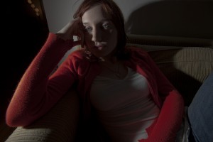 MATTHEW SWARTS A PERSON WITH LIGHT SKIN AND REDDISH HAIR SITS ON A COUCH, WEARING A RED CARDIGAN AND WHITE TOP, RESTING THEIR HEAD ON THEIR HAND AND GAZING THOUGHTFULLY TO THE SIDE IN DIM LIGHTING, EVOKING THE MOOD OF AN ART PIECE BY MATTHEW SWARTS.