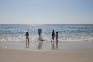 MATTHEW SWARTS A FAMILY PLAYS AT THE OCEANS EDGE ON A SUNNY BEACH. AN ADULT HOLDS A SMALL CHILD WHILE WAVES SPLASH AROUND THEM, AS THREE OTHER CHILDREN WADE IN THE SHALLOW SURF UNDER A CLEAR BLUE SKY.